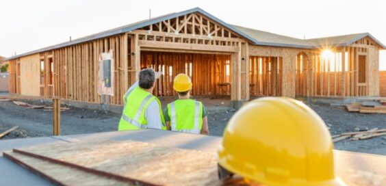 Two people wearing yellow hard hats and yellow safety vests are standing next to a home construction site.