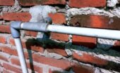 Exposed gray and white piping crosses an aged red brick wall with uneven mortar, cracks, and cement repairs.