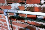 Exposed gray and white piping crosses an aged red brick wall with uneven mortar, cracks, and cement repairs.