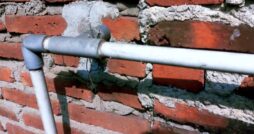 Exposed gray and white piping crosses an aged red brick wall with uneven mortar, cracks, and cement repairs.