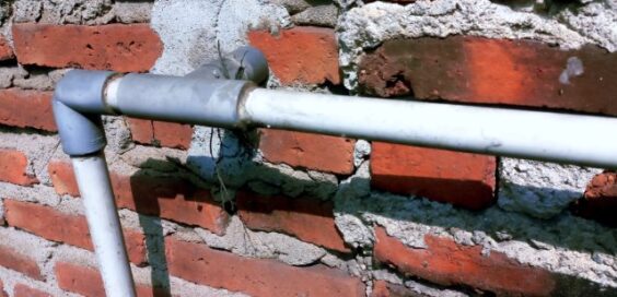 Exposed gray and white piping crosses an aged red brick wall with uneven mortar, cracks, and cement repairs.