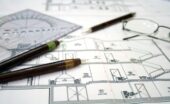 A close-up of the architectural design plans for a home with multiple pens, measuring tools, and eyeglasses on it.