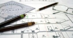 A close-up of the architectural design plans for a home with multiple pens, measuring tools, and eyeglasses on it.