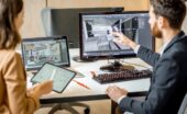 Designer points to a 3D room model on a desktop monitor while a colleague reviews plans on a tablet nearby.