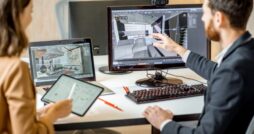 Designer points to a 3D room model on a desktop monitor while a colleague reviews plans on a tablet nearby.