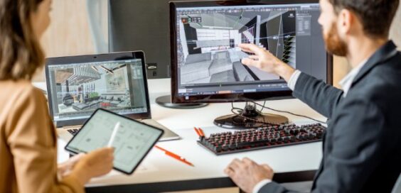 Designer points to a 3D room model on a desktop monitor while a colleague reviews plans on a tablet nearby.