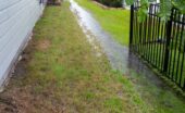 Excess rainfall traveling along the swale in the grass between two homes on a stormy day. One home has a black fence.