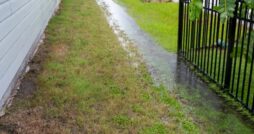 Excess rainfall traveling along the swale in the grass between two homes on a stormy day. One home has a black fence.
