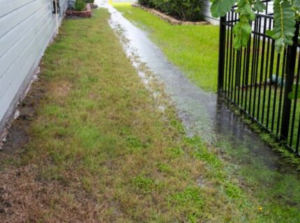 Excess rainfall traveling along the swale in the grass between two homes on a stormy day. One home has a black fence.