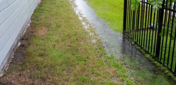 Excess rainfall traveling along the swale in the grass between two homes on a stormy day. One home has a black fence.