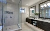 A gray bathroom with a white freestanding tub, glass shower, double sink vanity, large window, and tiled floor.