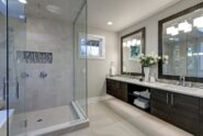 A gray bathroom with a white freestanding tub, glass shower, double sink vanity, large window, and tiled floor.