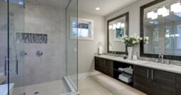 A gray bathroom with a white freestanding tub, glass shower, double sink vanity, large window, and tiled floor.