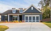 A gray ranch-style house with white trim, a two-car garage, brick accents, lawn, trees, and a concrete driveway.