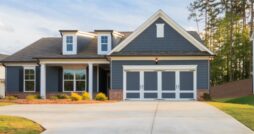 A gray ranch-style house with white trim, a two-car garage, brick accents, lawn, trees, and a concrete driveway.