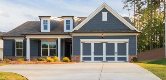 A gray ranch-style house with white trim, a two-car garage, brick accents, lawn, trees, and a concrete driveway.