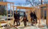 Five workers raising wooden wall frames at a residential building site, with lumber and tools scattered nearby.