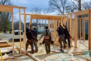 Five workers raising wooden wall frames at a residential building site, with lumber and tools scattered nearby.
