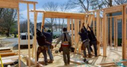 Five workers raising wooden wall frames at a residential building site, with lumber and tools scattered nearby.
