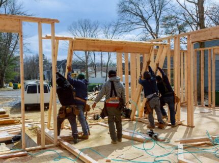 Five workers raising wooden wall frames at a residential building site, with lumber and tools scattered nearby.