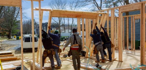 Five workers raising wooden wall frames at a residential building site, with lumber and tools scattered nearby.
