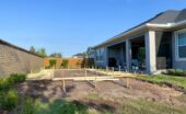 A backyard construction site with wooden stakes and framing laid out on excavated soil beside a house and covered patio.