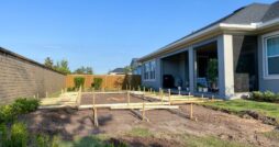 A backyard construction site with wooden stakes and framing laid out on excavated soil beside a house and covered patio.