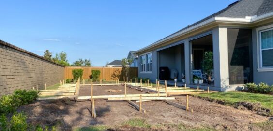 A backyard construction site with wooden stakes and framing laid out on excavated soil beside a house and covered patio.