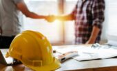 A yellow construction hard hat rests on a desk with papers and a level as two people shake hands in the background.