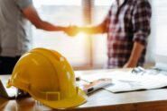 A yellow construction hard hat rests on a desk with papers and a level as two people shake hands in the background.