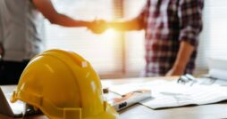 A yellow construction hard hat rests on a desk with papers and a level as two people shake hands in the background.