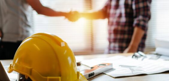 A yellow construction hard hat rests on a desk with papers and a level as two people shake hands in the background.