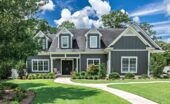A large dark gray house with white trim, dormer windows, a covered porch, and a curved walkway across a green lawn.