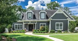 A large dark gray house with white trim, dormer windows, a covered porch, and a curved walkway across a green lawn.