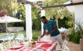 A man wearing a blue shirt placing glasses on an outdoor table for six with a plaid tablecloth under a pergola.