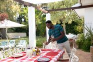 A man wearing a blue shirt placing glasses on an outdoor table for six with a plaid tablecloth under a pergola.