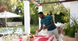 A man wearing a blue shirt placing glasses on an outdoor table for six with a plaid tablecloth under a pergola.