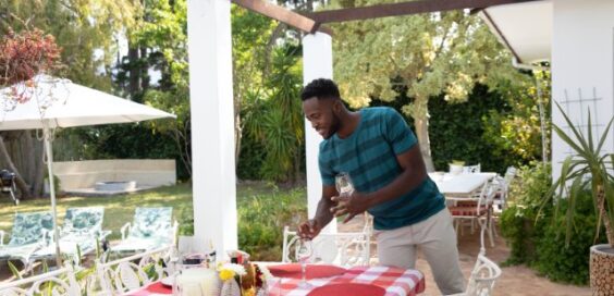 A man wearing a blue shirt placing glasses on an outdoor table for six with a plaid tablecloth under a pergola.
