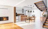 Modern open-plan home with a wood and metal staircase, white brick fireplace, wooden dining table, and bright kitchen.