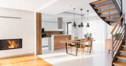 Modern open-plan home with a wood and metal staircase, white brick fireplace, wooden dining table, and bright kitchen.